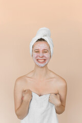 Young woman with problem skin with white care mask on her face. Girl with acne in bath towel on a beige background.