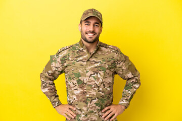 Military man isolated on yellow background posing with arms at hip and smiling