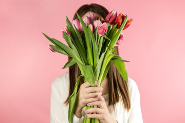 Portrait of a stylish young woman hiding her face with a bouquet of red tulips on a pink background.