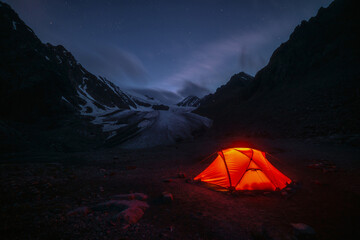 Awesome mountain landscape with vivid orange tent near large glacier tongue under clouds in night starry sky. Tent glow by orange light with view to glacier and mountains silhouettes in starry night.