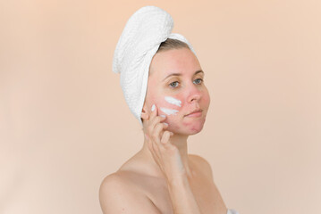 Young woman with problem skin with care cream on her face. Girl with acne in bath towel on a beige background.