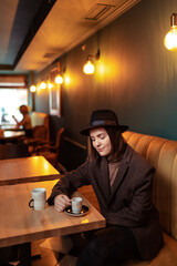 Drinking coffee. Pretty young woman in hat holding cup of beverage while sitting at cafe.