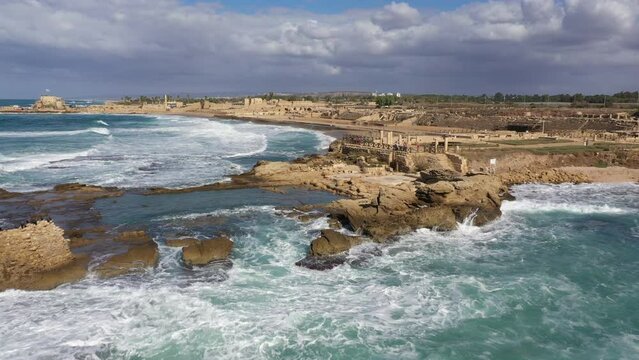 Revealing drone shot flying over rolling waves towards ancient Caesarea, excavated ruins of a Roman era city in Northern Israel
