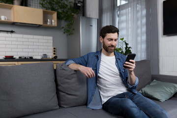 Thinking man looking at phone screen sitting on sofa at home