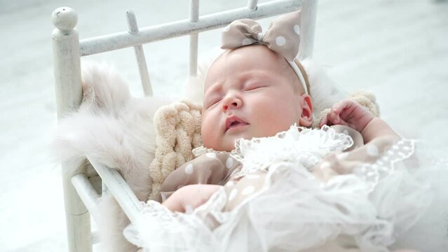 Adorable newborn girl in cute dress with dotted bow on head sleeps in small cot standing on white floor. Baby photo-shoot in studio close view