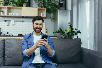 Happy man smiling and using phone sitting on sofa at home