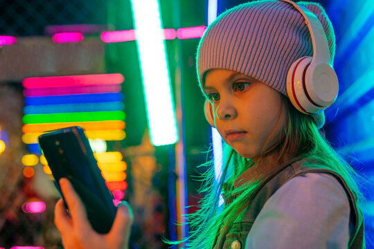 Fototapeta Little girl wearing headphones using smartphone on the colorful neon