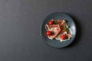 Prosciutto with tomatoes and fresh herbs. Delicacy on a dark plate on a dark tabletop.