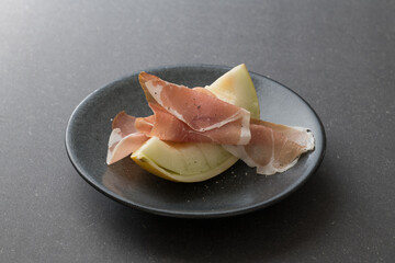 Sliced of Galia melon with iberico ham on a dark plate and black background.