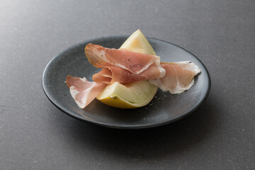 Sliced of Galia melon with iberico ham on a dark plate and black background.