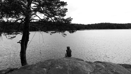 Black and white photo of a young woman in a sweatshirt and hood resting on a ridge overlooking a lake in northern Europe.