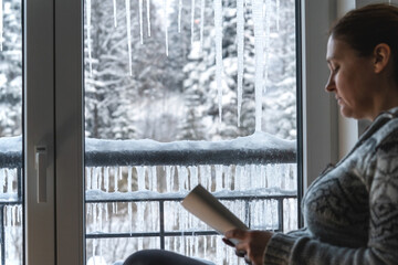 Snowy ice age.Icicles on the windows in focus.A woman sits by the window and reads.Very cold weather outside. 