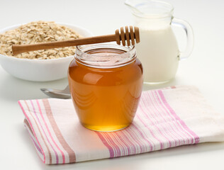 honey in a glass transparent jar and a wooden stick on a white table, behind a decanter with milk and oatmeal in a plate