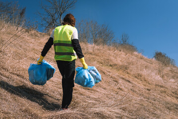 Man nature activist in yellow vest carries blue trash bags along the pathway in the sunny field