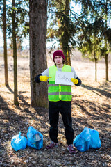 Male nature activist in green vest holds white paper with ecology word while standing near blue trash bags