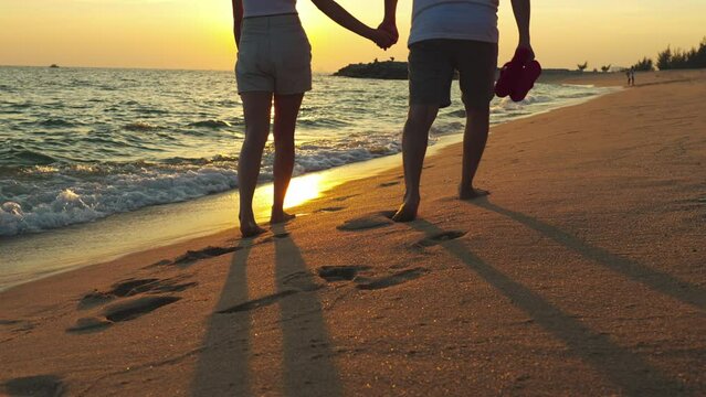 Couple walking together on the beach at sunset, young happy couple holding hands walking along beach, walking barefoot and carrying shoes, outdoor leisure time by the seaside.