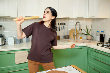 A young woman is fooling around and grimacing in the kitchen, singing into a rolling pin as if into...