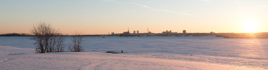 Panorama with a view of the snow-covered river and the city on the far bank at sunset.