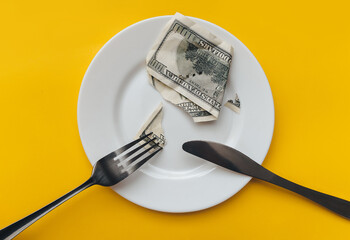 The cut dollar bill lies on a white plate, pierced by a fork near the knife on a yellow tablecloth. Business lunch, banking, rentier life and financial well-being concept.
