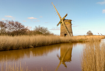 Derelict and abandoned Norfolk drainage Windmill, next to a waterway. With broken sails and open doorway. Reflections in water. Countryside. Landscape image with space for text. Near Thurne, England. - 493835677