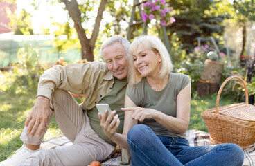 Naklejka premium Happy senior spouses sitting on blanket during picnic in garden and using smartphone, resting outdoors on spring day