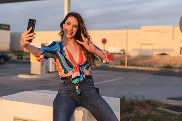 chica con camisa de colores y sonriendo sentada en la calle haciéndose selfies con una gasolinera...
