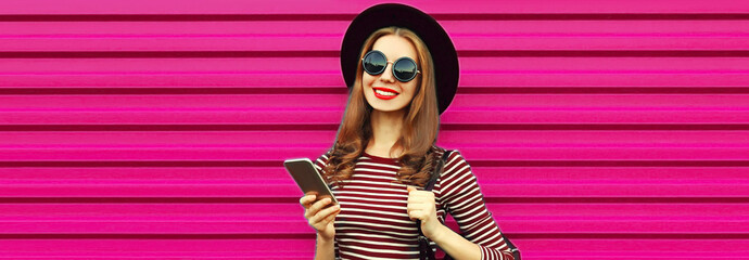 Portrait of happy smiling young woman with smartphone wearing black round hat on pink background