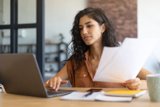 Focused young businesswoman working or studying on laptop, holding documents, sitting at desk indoors, copy space