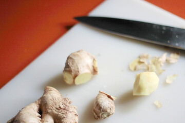 Ginger is being sliced on a plastic chopping board with a knife on an orange table.