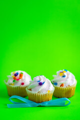 Three white frosting cupcakes with colorful sprinkles on bright green background and light blue bowtie.