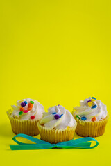Three white frosting, colorful sprinkles cupcakes with light blue bowtie on bright greenish yellow background.