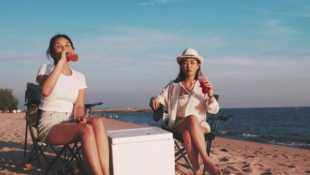 Two teenage girls sitting on beach clinking bottles and drinking soda, group of friends enjoying drinks and playing guitar in camping beach at sunset, vacation and lifestyle concept.