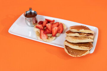 Close-up shot of pancakes, slices of banana and strawberry and hot chocolate on white plate shot with selective focus from opposite or side angle on isolated area, orange background.