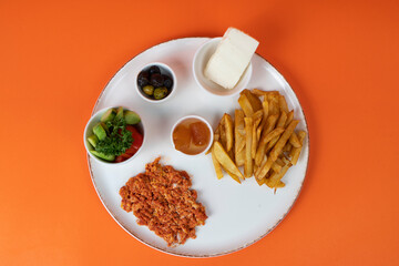 Top view of salad plate, french fries, citrus jam, olive platter, feta cheese and menemen on white plate, shot with selective focus from overhead angle on isolated area, orange background.
