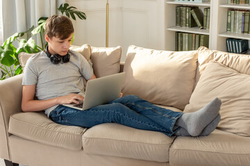 A teenager lying on a beige sofa, in a sunny room, wearing black headphones around his neck, looks...