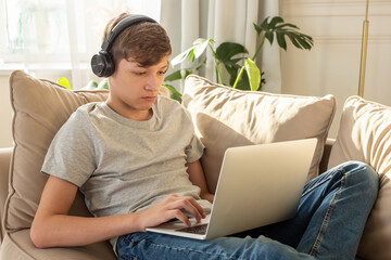 Portrait of teenage , sits on a sofa in a room, wearing black headphones on his head, using a laptop.