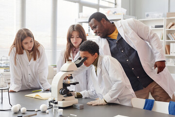 Side view portrait of young black girl looking into microscope with while doing experiments with group of children in school chemistry lab, copy space