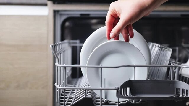Close-up of human hand puts or insert dirty plate to the basket of an open automatic stainless built-in dishwasher machine with dish utensils inside in modern home kitchen. Household domestic life