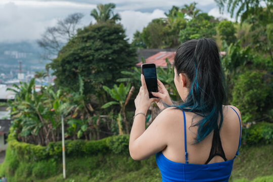 young latina woman with blue hair taking a picture of a landscape in the beautiful colombian mountains. influencer girl creating content for her social networks, while hiking.