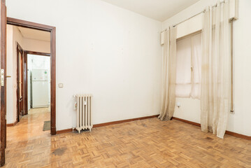Empty living room with oak parquet flooring, cast iron radiator and window with white curtains