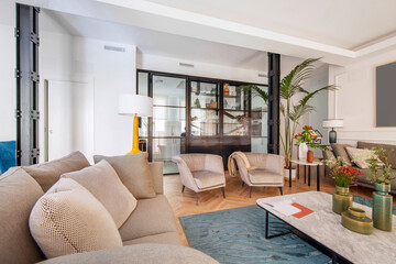 Living room with beautiful contemporary decor with light brown fabric sofa, armchairs, vases, marble table and steel and glass kitchen wall