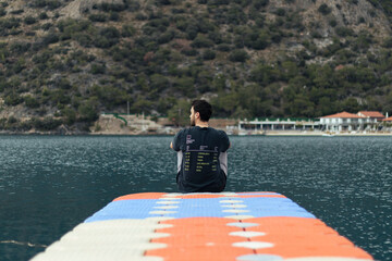 Fototapeta premium Young man sitting alone on dock. He is watching sea view. Her face is not seen in photo. Unrecognizable person. Quiet place.