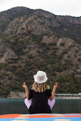 Back view of young woman with hat practices yoga and meditation on dock. Sea background. Healthy life and fresh air concept. Vertical banner.	