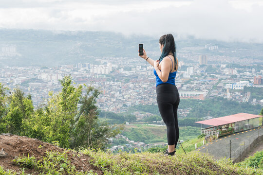 young woman. latin influencer recording a video for her social networks on top of a mountain, in the background the city of Pereira-Colombia. girl with fitnes clothes hiking on top of a hill.