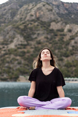 Fototapeta premium Cross-legged young woman. Stretching young girl on colorful dock by sea. Sport, healthy life, meditation and mindfulness. Beautiful landscape. Fresh air concept.