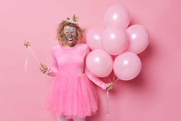 People and holidays concept. Positive optimistic young woman with claymask on face excaims loudly hass fun on bachelorette party wears festive clothes holds bunch of inflated helium balloons