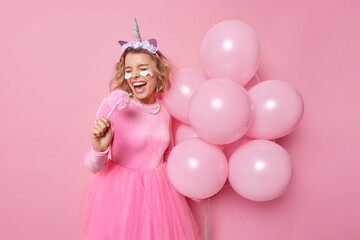 Obraz na płótnie Canvas Energetic positive woman sings and foolishes around poses with bunch of inflated balloons wears dress applies beauty patches under eyes isolated over pink background. Festive occasion concept