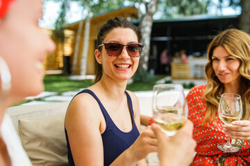 Three adult beautiful caucasian women female friends sitting outdoor in backyard with glasses of white wine having fun celebrating and drinking in summer day on vacation holiday - real people leisure