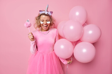 Obraz na płótnie Canvas Joyful glad woman wears dress unicorn headband applies beauty patches holds bunch of inflated balloons has fun on party celebrates special occasion poses against pink background. Celebration