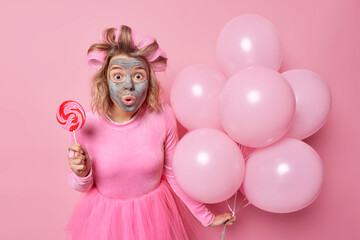 Impressed stunned woman stares bugged eyes has shocked expression wears hair rollers beauty mask festive dress celebrattes something poses with bunch of balloons sweet lollipop isolated on pink wall
